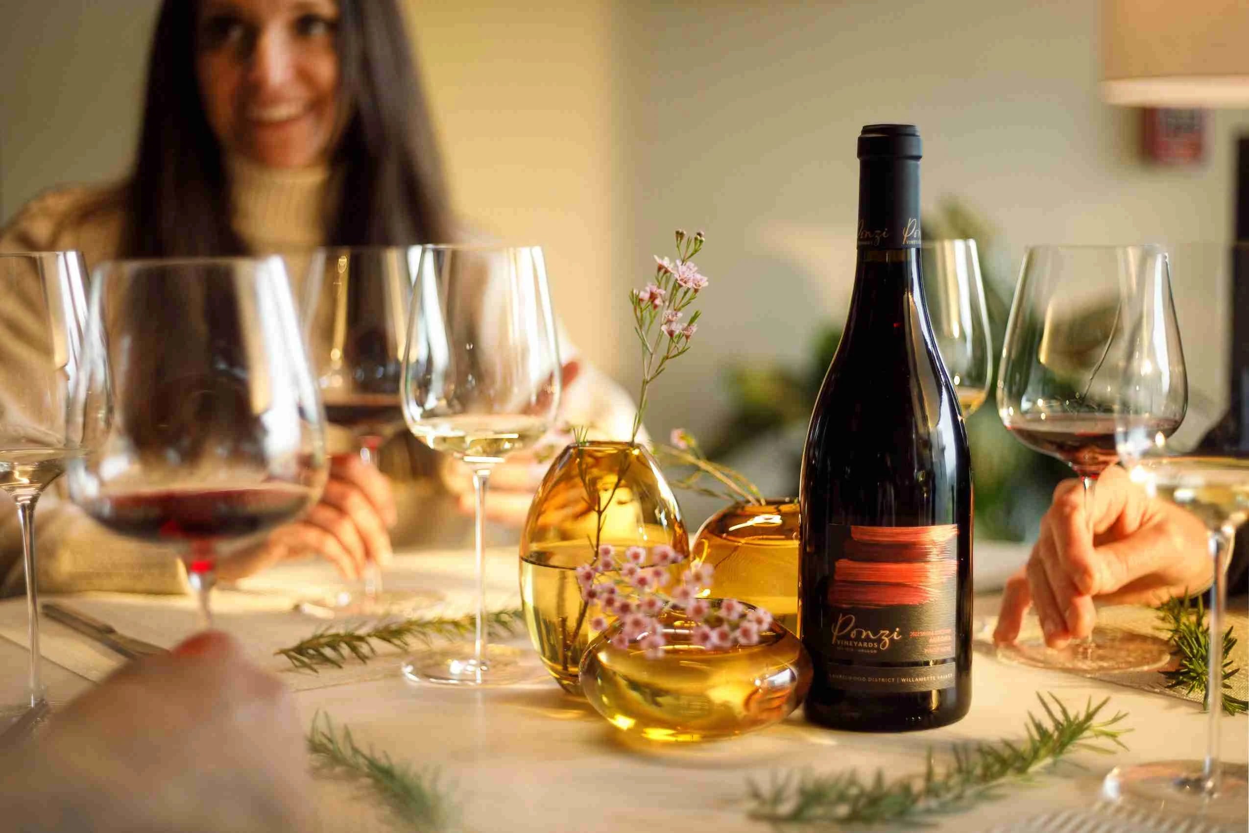 A table filled with wine glasses, small flower vases, and a Ponzi Vineyards wine bottle. A woman is blurred in the background.